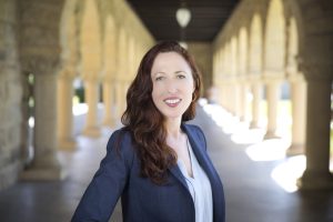 Photo of Garance Burke, a white woman with long brown hair wearing a blue blazer and standing outside, with series of columns descending behind her in the background