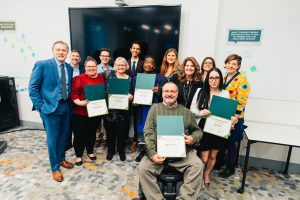 A group of thirteen people stand in a semi-circle, with five adults in the front row holding certificates of completion.