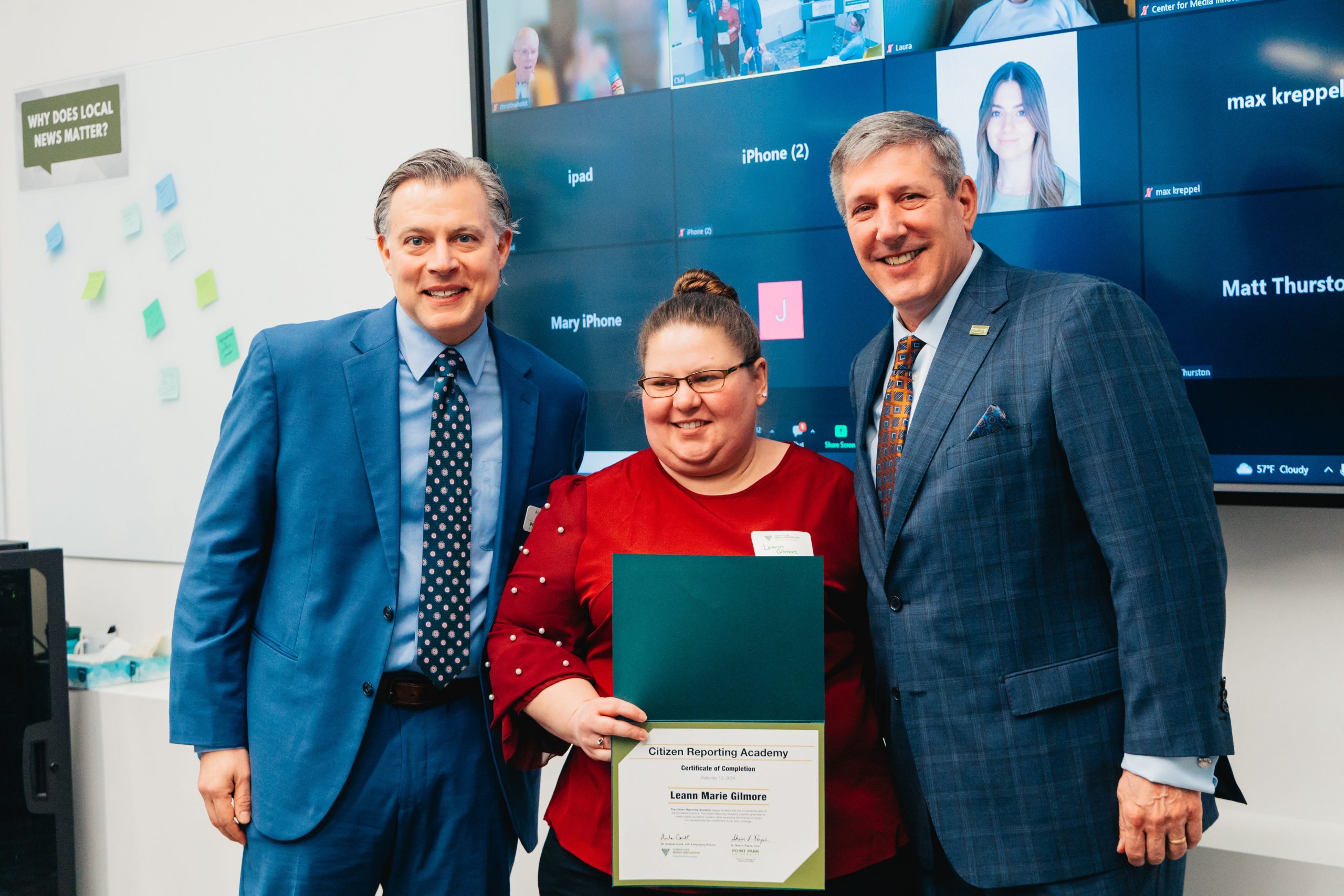 Two men in suits stand on either side of a woman holding a certificate of completion.