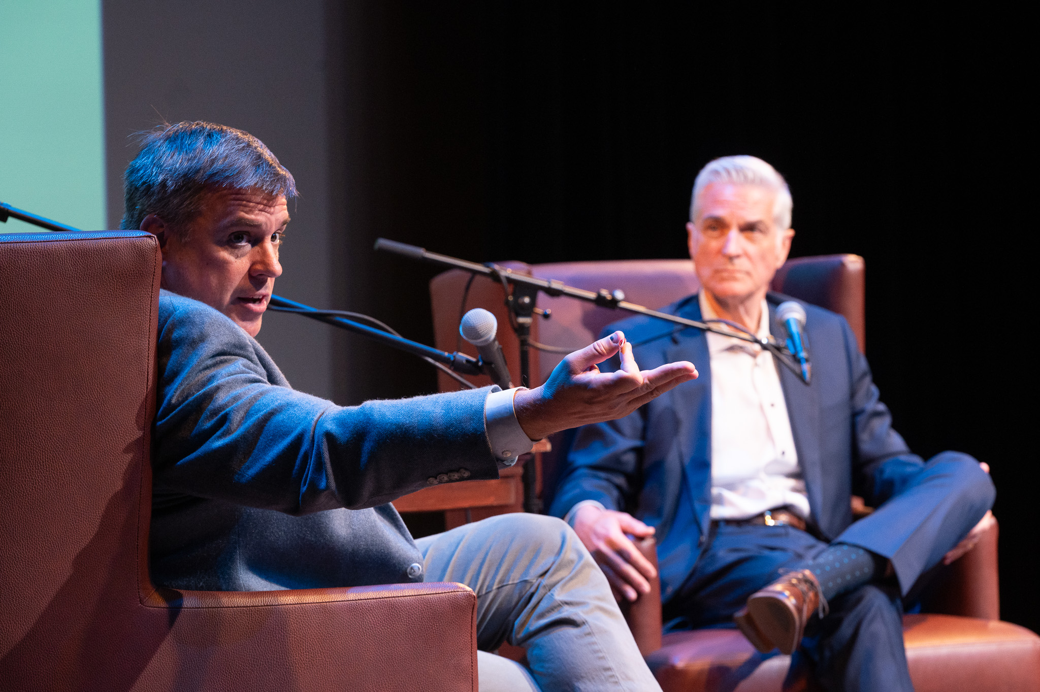 Image of two white men sitting in leather chairs conversing before an audience.