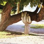 Photo of a cement sculpture of a hand placed under a tree branch that is growing low and horizontal to the ground.
